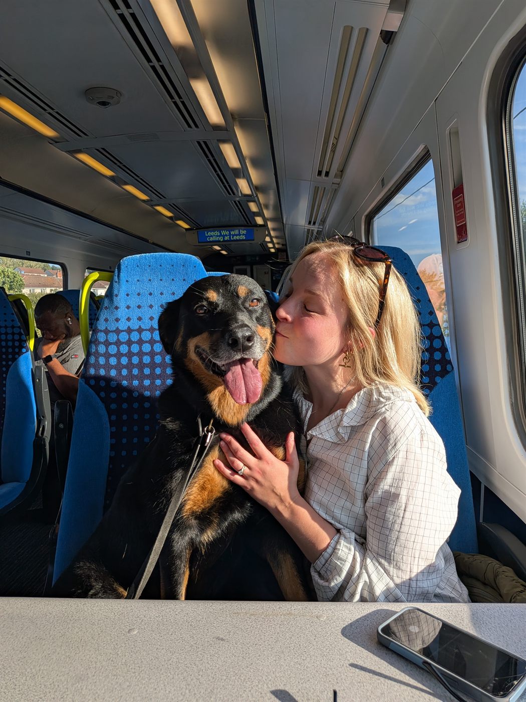 Joanna on a train with her eyes closed, kissing her dog Queenie on the cheek. Queenie is grinning at the camera with her tongue out.